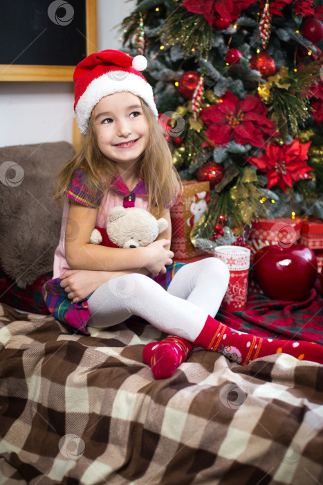 Скачать Маленькая девочка, держащая в руках плюшевого мишку, сидит на клетчатом одеяле в рождественских украшениях возле рождественской елки с коробками подарков и шляпой Санты. Новый год, детская игра фотосток Ozero