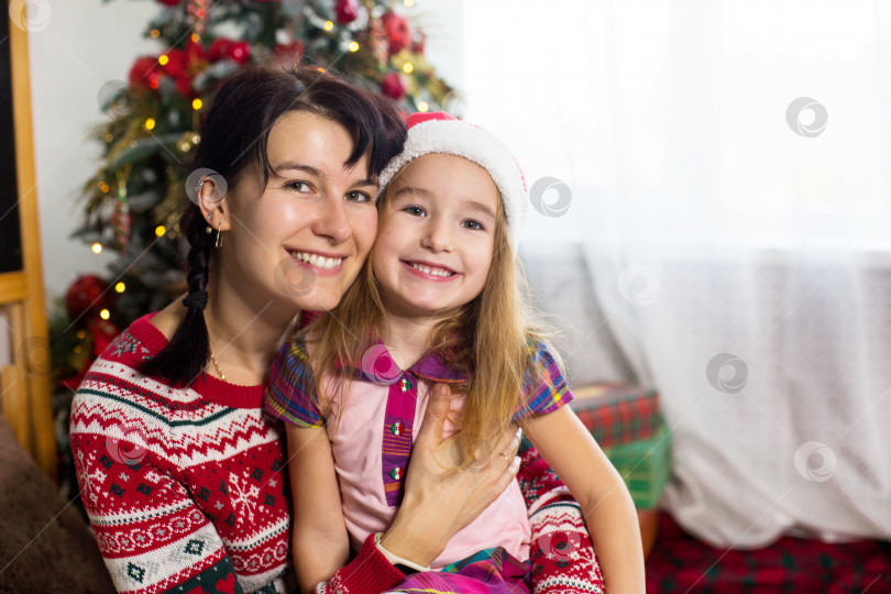 Скачать Мама и дочь обнимаются, счастливо улыбаются и смотрят в камеру, сидят дома возле рождественской елки в удобной одежде и шляпе Санты. Рождество, канун нового года дома. Декор, подарочные коробки, копировальное пространство фотосток Ozero
