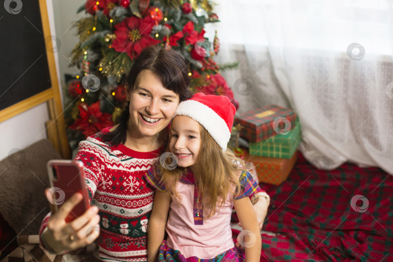 Скачать Мама и дочь сидят возле рождественской елки, обнимаются, делают фотографии и селфи на телефон, общаются по видеосвязи, веселятся. Рождество, канун нового года дома. Пространство для копирования фотосток Ozero