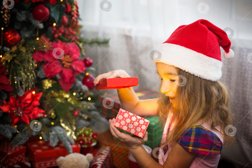 Скачать Маленькая девочка в шляпе Санты открывает красную коробку с подарком и золотым волшебным огоньком возле рождественской елки. Праздничный декор, пуансеттии на елках, Новый год. Радость, удивление, детские эмоции. Копировальное пространство фотосток Ozero