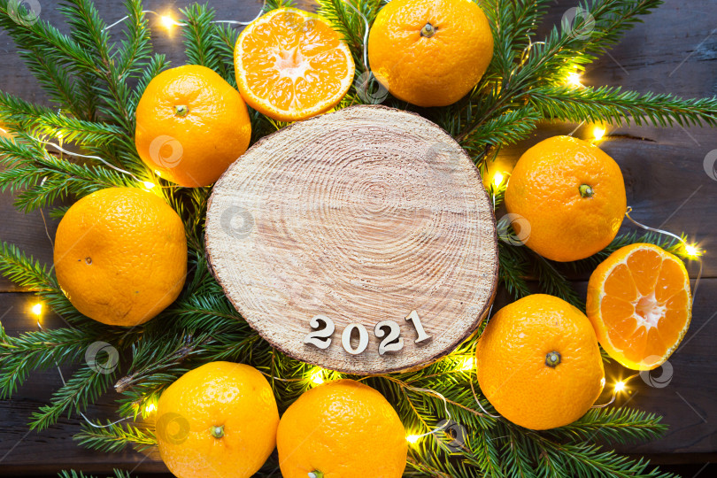 Скачать Новогодний праздничный фон на круглом срезе дерева, окруженном мандаринами, живыми еловыми ветками и гирляндами из золотых огней, с деревянными цифрами, датированными 2021 годом. Цитрусовый аромат, Рождество. Пространство для текста. фотосток Ozero