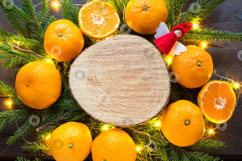 Скачать Новогодний праздничный фон на круглом срезе дерева, окруженном мандаринами, живыми еловыми ветками и гирляндами из золотых огней, с деревянным пространством для текста. Цитрусовый аромат, ломтики апельсина, Рождество. рамка фотосток Ozero