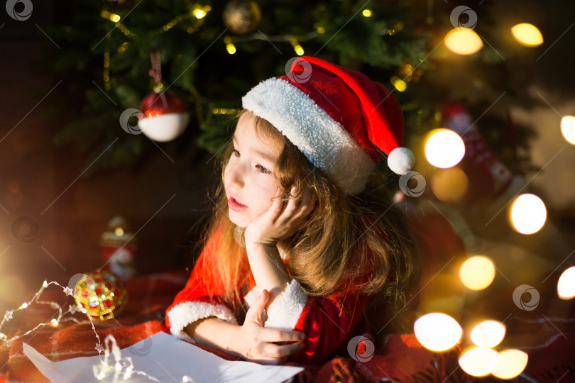 Скачать Маленькая девочка в шляпе Санты и красном платье под рождественской елкой мечтает, ожидая праздника, лежа на клетчатом одеяле. Письмо на клочке бумаги, подарки. Новый год, Рождество. расфокусировка огней Маленькая девочка в шляпе Санта-Клауса и красном платье под рождественской елкой мечтает, ожидая праздника, лежа на клетчатом одеяле. Письмо на клочке бумаги, подарки. Новый год, Рождество. расфокусировка света фотосток Ozero