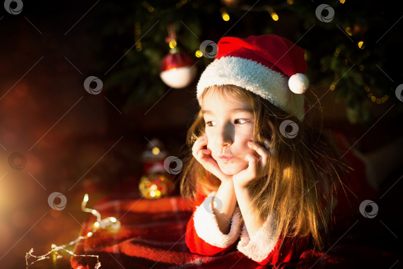 Скачать Маленькая девочка в шляпе Санты и красном платье под рождественской елкой мечтает, ожидая праздника, лежа на клетчатом одеяле. Письмо на листе бумаги, подарки. Новый год, Рождество. расфокусировка света фотосток Ozero