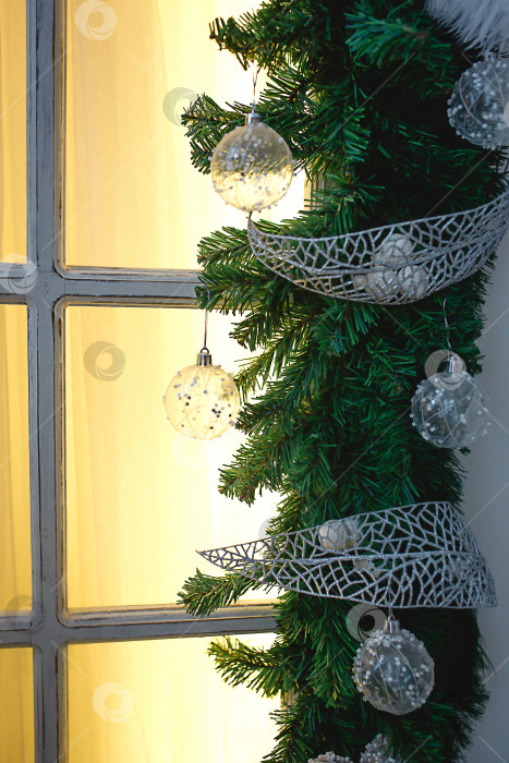 Скачать Рождественские украшения из еловых веток, белых прозрачных шаров и лент обрамляют окно с деревянной рамой и желтым светом. Новогодняя праздничная атмосфера, домашний уют. Пространство для текста фотосток Ozero