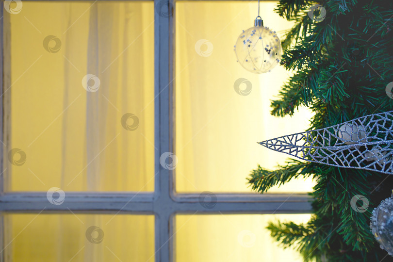 Скачать Рождественские украшения из еловых веток, белых прозрачных шаров и лент обрамляют окно с деревянной рамой и желтым светом. Новогодняя праздничная атмосфера, домашний уют. Пространство для текста фотосток Ozero