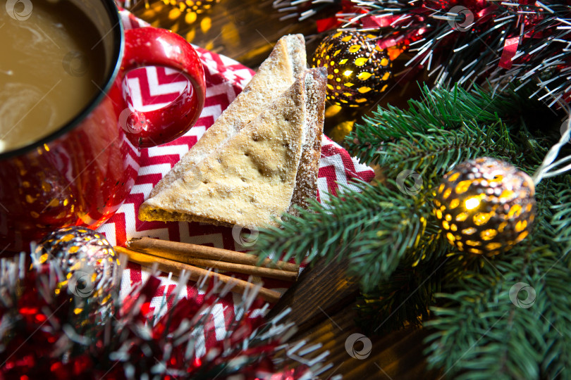 Скачать Треугольное печенье с сахарной пудрой и корицей на салфетке с зигзагообразным рисунком в рождественском декоре. Красная кофейная кружка с зефиром, еловыми ветками, гирляндой, новый год, уют. фотосток Ozero