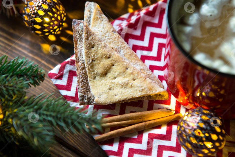 Скачать Треугольное печенье с сахарной пудрой и корицей на салфетке с зигзагообразным рисунком в рождественском декоре. Красная кофейная кружка с зефиром, еловыми ветками, гирляндой, новый год, уют. фотосток Ozero