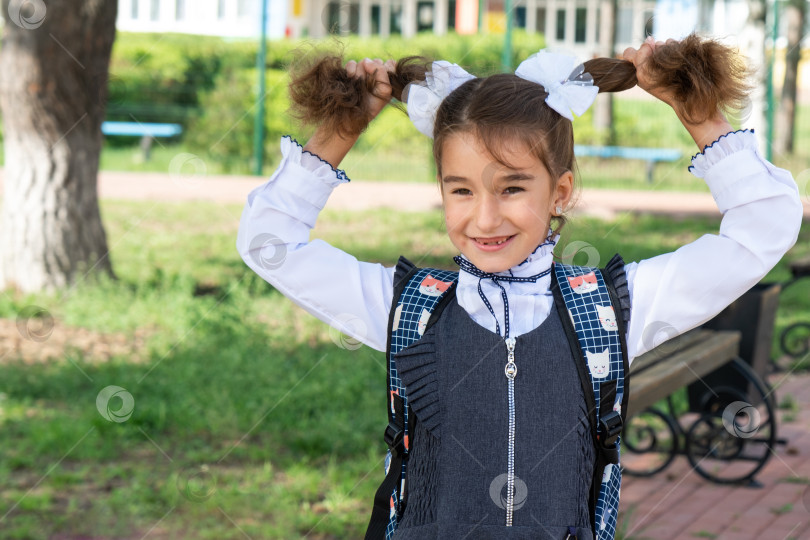 Скачать Веселая забавная девочка с беззубой улыбкой в школьной форме с белыми бантами на школьном дворе. Возвращение в школу, 1 сентября. Счастливый ученик с рюкзаком. Начальное образование, начальный класс. фотосток Ozero