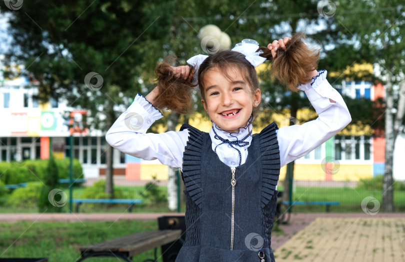 Скачать Веселая забавная девочка с беззубой улыбкой в школьной форме с белыми бантами на школьном дворе. Возвращаюсь в школу, 1 сентября. Счастливый ученик. Начальное образование, начальный класс. Портрет студента фотосток Ozero