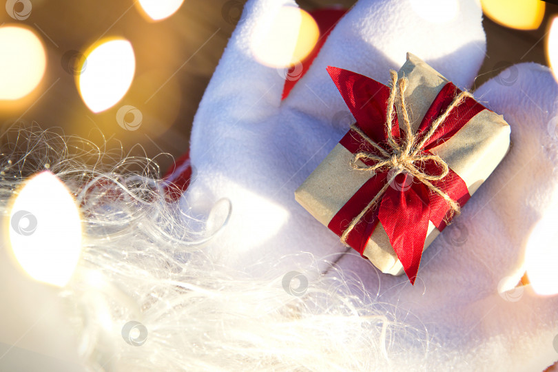 Скачать Коробка с рождественским подарком в руках Деда Мороза в белых варежках. Красный костюм, борода, гирлянды огней - все как в тумане. Новый год, подготовка, ожидание чуда, сбывшейся мечты. Закрыть фотосток Ozero
