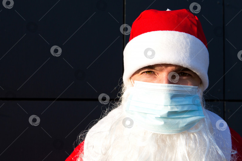 Скачать Санта-Клаус с белой бородой, в красном пальто и медицинской маске. Рождество в условиях пандемии коронавируса, сезонных заболеваний, ОРВИ и пневмонии в праздничные дни. Защита от вирусов в новом году фотосток Ozero