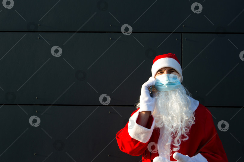 Скачать Санта-Клаус в медицинской маске со смартфоном. Онлайн-поздравления, заказ услуг на Рождество и новый год. Социальная дистанция в условиях эпидемии коронавируса - новая реальность. Пространство для копирования фотосток Ozero