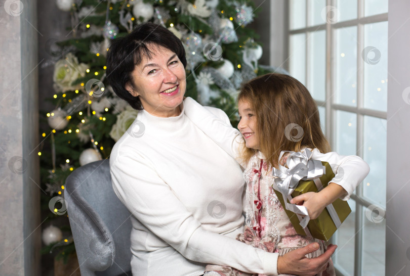 Скачать Бабушка с маленькой девочкой на фоне рождественских украшений и большого окна. Семейный праздник, эмоции, подарочная коробка. Внучка на коленях у бабушки. новый год фотосток Ozero