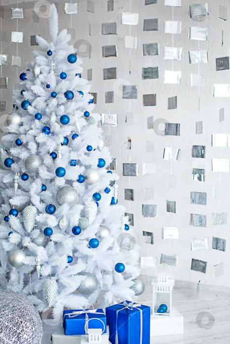 Скачать Рождественская белая искусственная елка в светло-серой комнате украшена голубыми и серебристыми воздушными шарами. Коробки с подарками, украшения из кубиков на стене. новый год. Пространство для текста, холодный фон фотосток Ozero