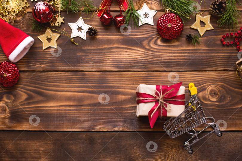 Скачать Рождественская плоская обложка на тему покупок, тележка с подарочной коробкой. Покупка подарков на новый год. Черная пятница, распродажа, декор из звезд, шара, бусин, гирлянд на деревянном фоне. Пространство для текста фотосток Ozero