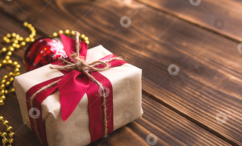 Скачать Коробка с рождественским подарком из светлой бумаги перевязана красной лентой на деревянном фоне с декором. Подвеска на рождественскую елку, бусы, гирлянда. Новый год. место для текста фотосток Ozero