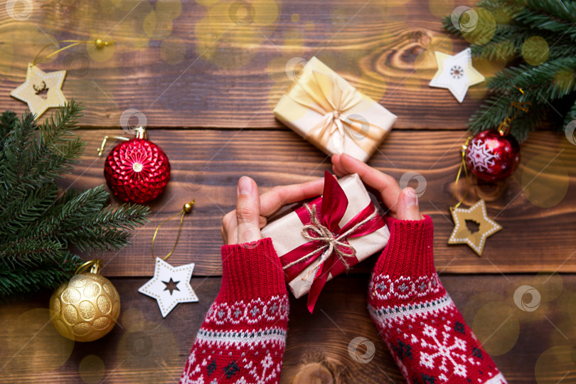 Скачать Женские руки в вязаном свитере с орнаментом держат в ладонях коробку с рождественским подарком на столе среди декора. Новый год, упаковка и приготовление подарков, еловые ветки, украшение фотосток Ozero