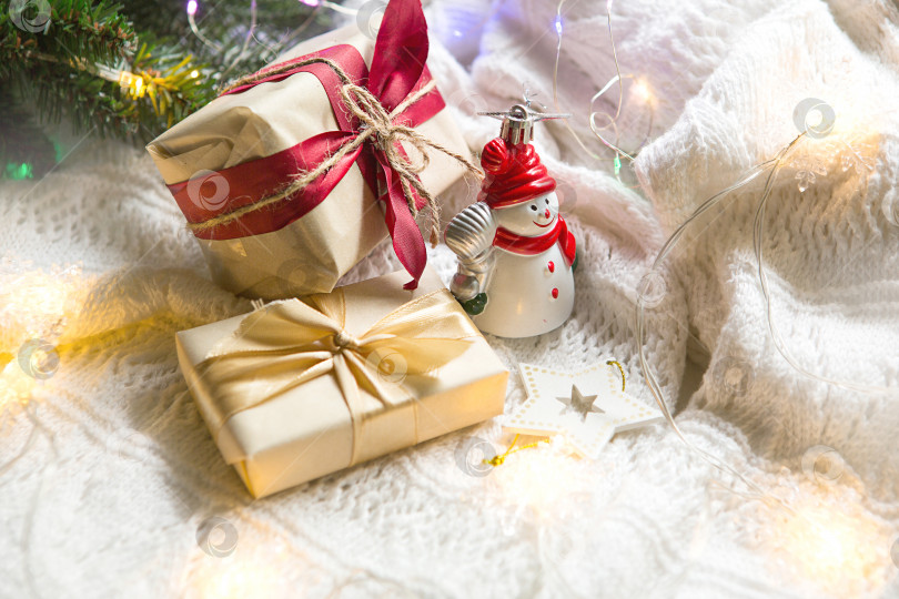 Скачать Коробки с рождественскими подарками и украшениями снеговика на белом вязаном одеяле в огнях гирлянд и еловых веток. Новый год, праздничная атмосфера. Пространство для текста фотосток Ozero