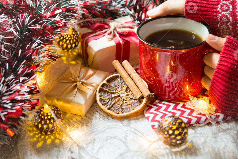 Скачать Женская рука в теплом свитере держит красную кружку с горячим напитком на столе с рождественскими украшениями. Новогодняя атмосфера, палочки корицы и ломтик сушеного апельсина, подарки, гирлянда и мишура фотосток Ozero