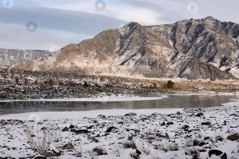 Скачать Скалистый заснеженный берег красивой горной реки, протекающей по живописной зимней долине солнечным декабрьским днем. фотосток Ozero