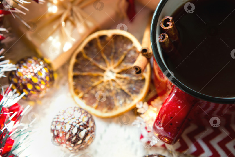 Скачать Красная кружка с чаем и палочками корицы, ломтик сушеного апельсина, рождественский декор, подарочные коробки и гирлянды на столе. Лежал плашмя. новый год фотосток Ozero