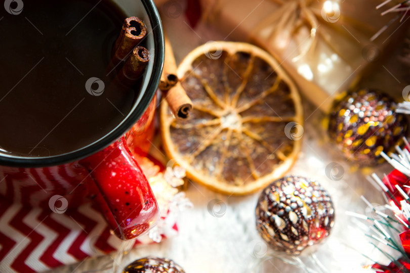 Скачать Красная кружка с чаем и палочками корицы, ломтик сушеного апельсина, рождественский декор, подарочные коробки и гирлянды на столе. Лежал плашмя. новый год фотосток Ozero
