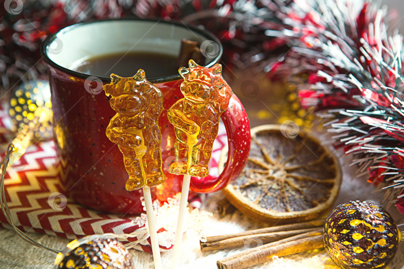 Скачать Два леденца на палочке в форме символа нового 2021 года - быка на фоне рождественского декора с красной чашкой чая. Гирлянды, корица и сушеный апельсин - атмосфера тепла, уюта и волшебства. фотосток Ozero