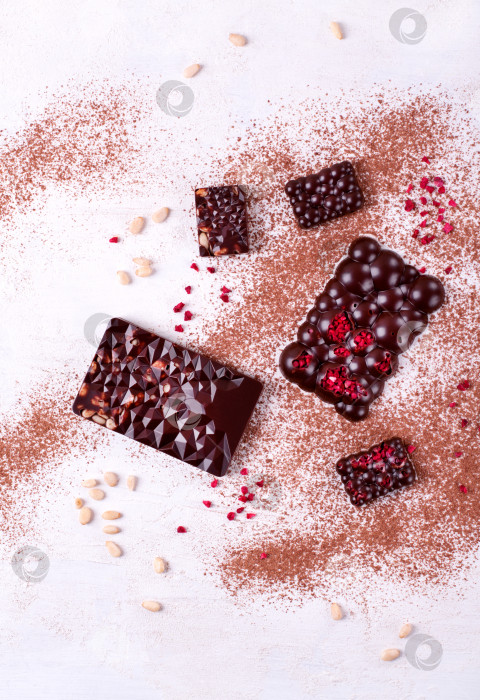 Скачать Шоколадные батончики разных размеров и форм с сублимированными ягодами и кедровыми орешками на белом столе с какао-порошком. фотосток Ozero