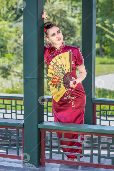 Скачать Молодая азиатская девушка прогуливается в национальной одежде с веером по парку. Китаянка в красном традиционном платье, портрет крупным планом фотосток Ozero