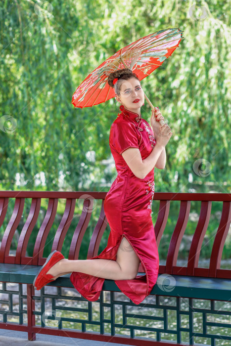 Скачать Молодая азиатская девушка прогуливается в национальной одежде с красным зонтиком по парку. Китаянка в красном традиционном платье, портрет крупным планом фотосток Ozero