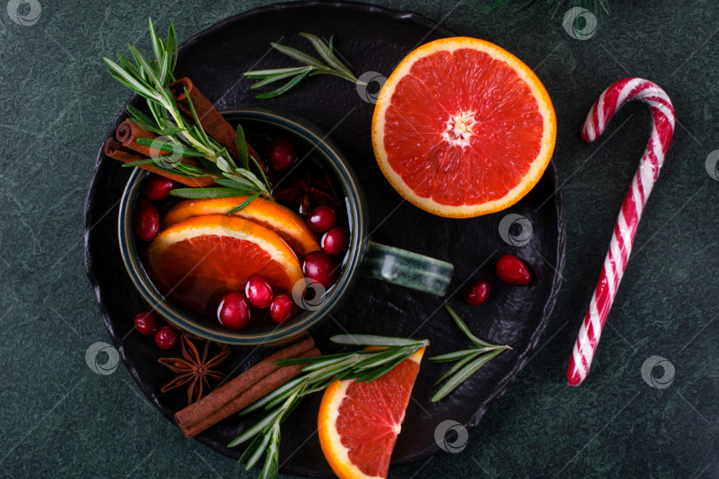 Скачать Глинтвейн подается с красным апельсином, корицей, клюквой и розмарином в зеленой керамической кружке на темном столе. Согревающий напиток на Рождество и зиму. фотосток Ozero