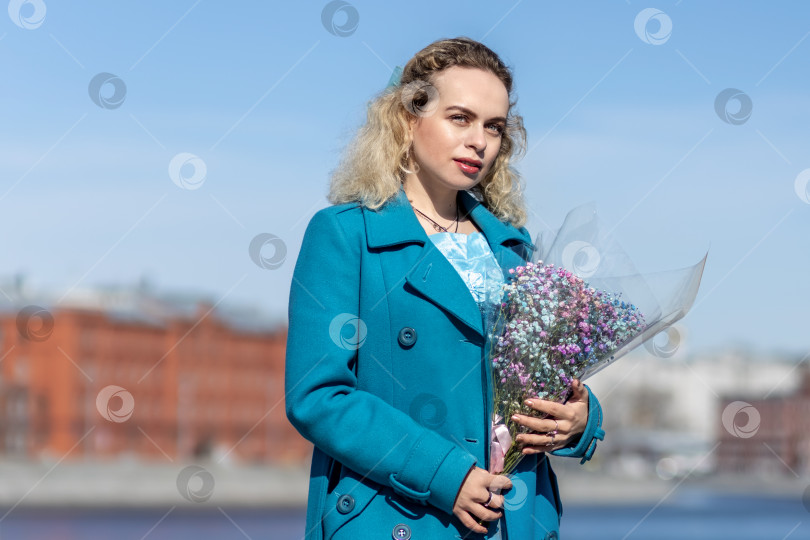 Скачать Молодая блондинка в синем пальто стоит с букетом разноцветных гипсофил на набережной в парке. Концепция весеннего праздника - 8 марта, Пасха, Женский день. фотосток Ozero