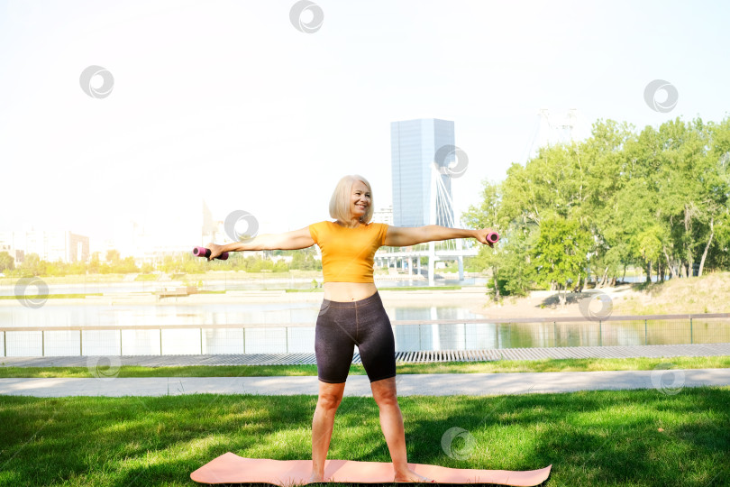 Скачать Пожилая женщина занимается спортивными упражнениями с гантелями в летнем парке. Концепция активного образа жизни. фотосток Ozero