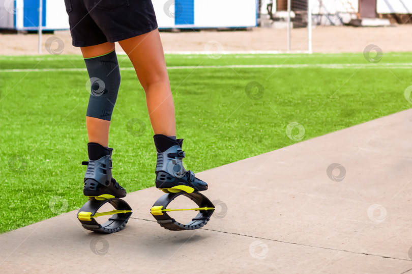 Скачать Молодая женщина в специальной обуви для фитнеса выполняет упражнения для восстановления после травм. Повязка на колене. Зеленое поле с искусственным покрытием на стадионе. фотосток Ozero