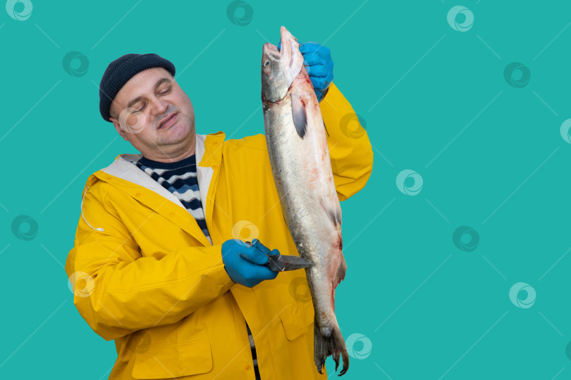 Скачать ловец очищает чешую пойманной рыбы. мужчина в рыбацком костюме на фоне моря. рыболов держит в руках нож. работа рыбака. фотосток Ozero