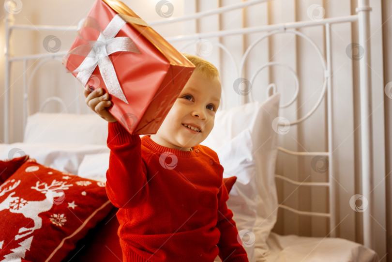 Скачать Улыбающийся забавный ребенок в красном вязаном свитере сидит на кровати и трясет рождественским подарком в руке. Рождественская концепция. фотосток Ozero