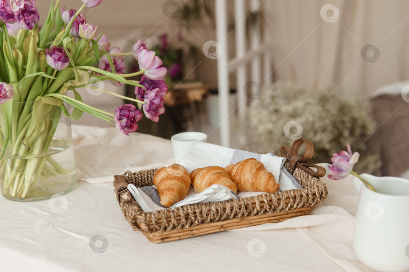 Скачать Свежие круассаны в плетеной корзинке на столе рядом с вазой с сиреневыми тюльпанами. фотосток Ozero