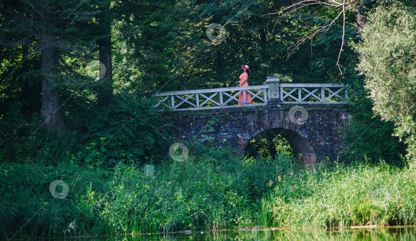 Скачать взрослая женщина в длинном коралловом платье и широкополой шляпе на старом мосту в лесу у пруда фотосток Ozero