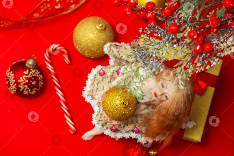 Скачать кукольная девочка с рождественским шаром в руках сидит под веткой рождественской елки среди новогодних украшений на красном фоне фотосток Ozero