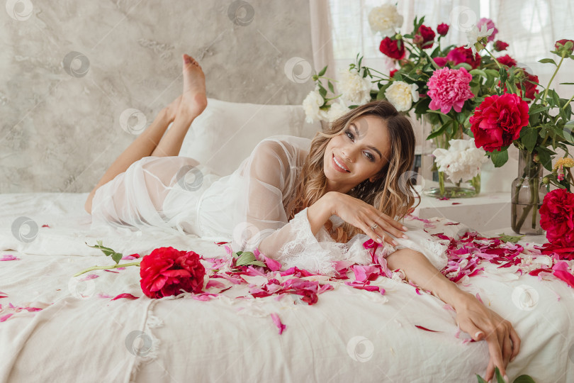 Скачать Красивая девушка в белой ночной рубашке дома, в окружении весенних цветов на большой кровати. Комната, украшенная букетами пионов фотосток Ozero