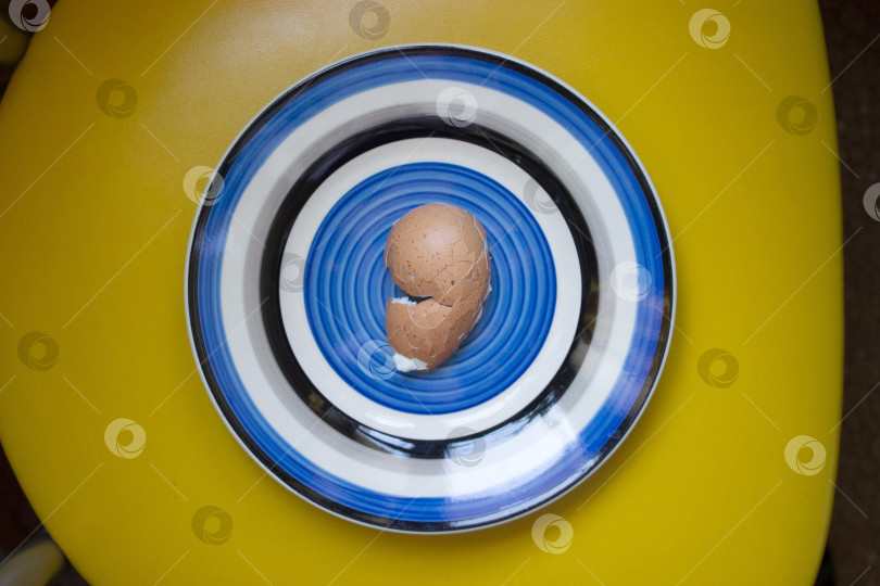Скачать Разбитая коричневая в крапинку яичная скорлупа на круглой синей тарелке на желтом фоне. Вареное куриное яйцо. Концептуальная основа фотосток Ozero