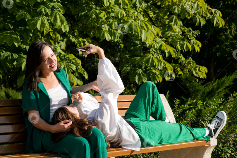 Скачать Мама и дочь-подросток на скамейке в парке смотрят на мобильный фотосток Ozero