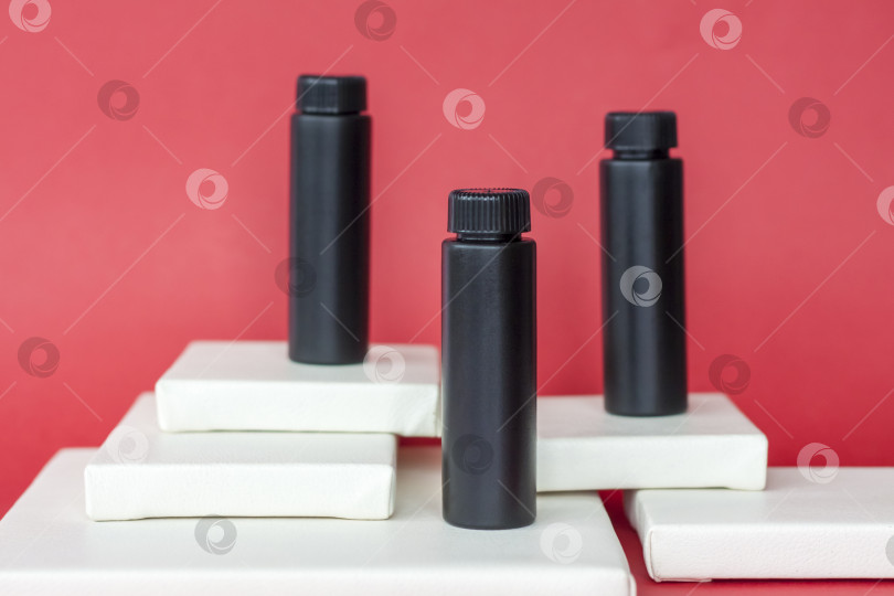 Скачать Черные бутылочки с кремом стоят на белых многоуровневых подставках. Черные бутылки на красном фоне. Уход за кожей, увлажняющий и питательный крем или солнцезащитный крем. фотосток Ozero