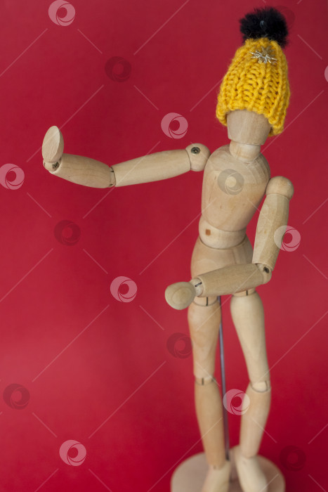 Скачать Деревянный манекен мужчины в вязаной желтой шапочке указывает на место рядом с ним. Манекен мужчины, сделанный из дерева на красном фоне. фотосток Ozero