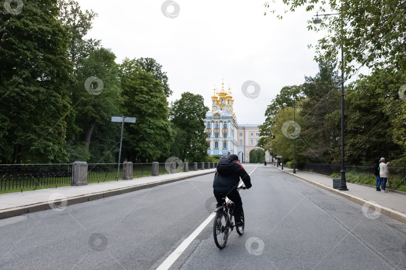 Скачать Велосипедист в чёрном на фоне Екатерининского дворца едет по дороге, зелёные деревья по бокам - Пушкин, санкт-Петербург, Россия, август 2023 фотосток Ozero