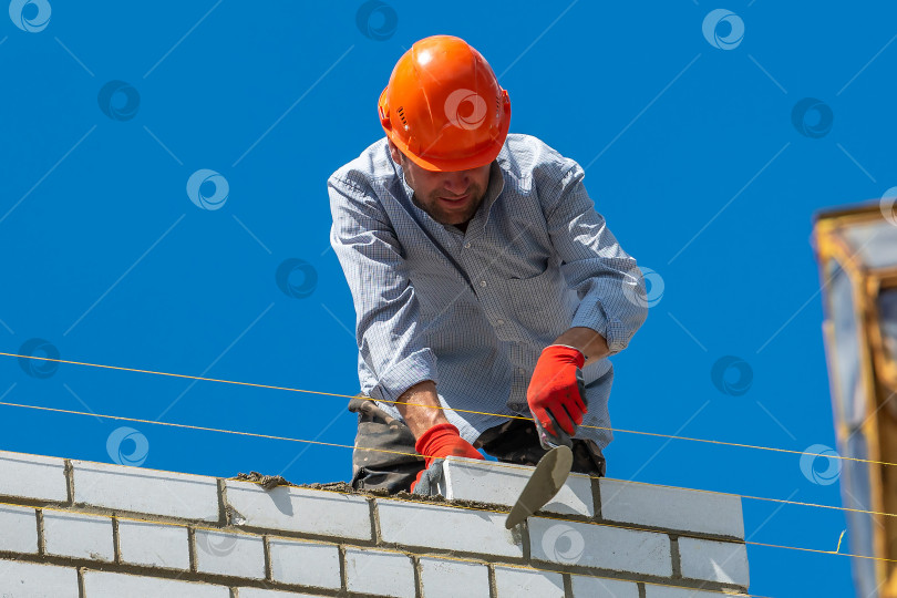 Скачать Череповец, Россия, 24 июня 2022 года рабочий-строитель возводит кирпичную стену на строительной площадке нового дома. Кирпичная кладка, строитель кладет кирпич. фотосток Ozero