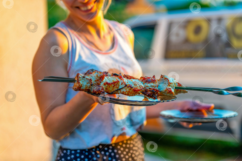 Скачать Сочные кусочки мяса на шампурах и в тарелке держит в руках молодая улыбающаяся девушка или женщина, не в фокусе. Приготовление шашлыка на природе на даче по выходным. фотосток Ozero