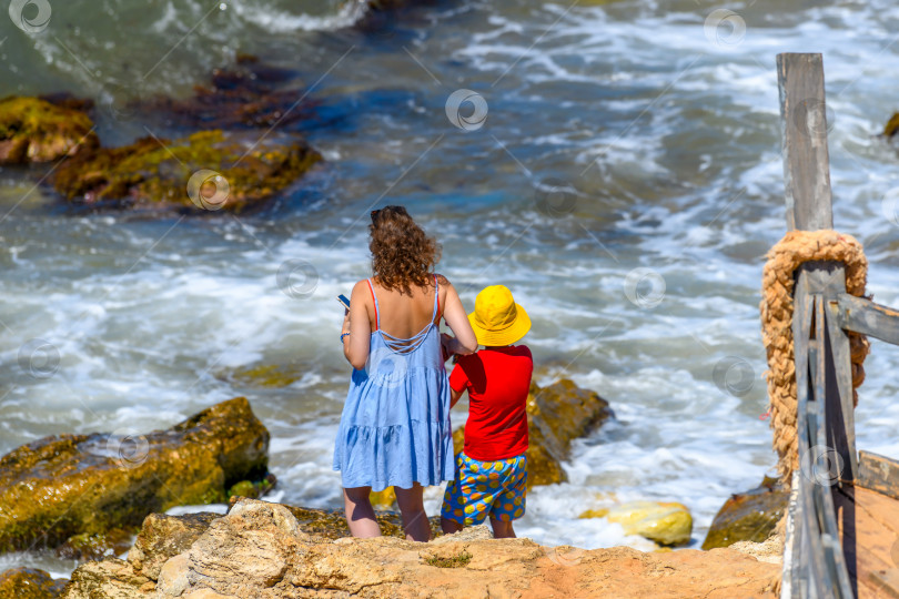 Скачать Мать со своим маленьким сыном стоят на скалистом морском берегу. Мама с ребенком, летние каникулы на море. фотосток Ozero