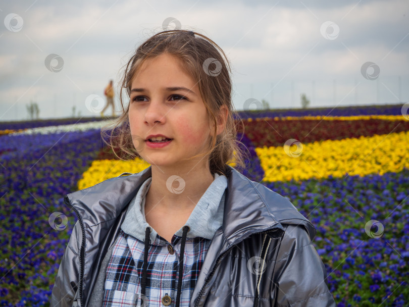 Скачать Красивая девушка смотрит вдаль, стоя на фоне разноцветной цветочной клумбы. Портрет девочки-подростка в сером жакете и клетчатой рубашке ранней весной на открытом воздухе фотосток Ozero